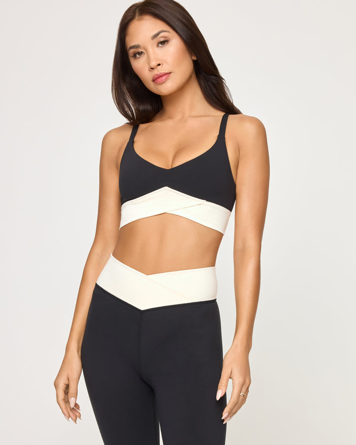 Close-up front view from the waist up of the LSPACE Unmatched Legging in black-cream on a model against a white background, highlighting the cream crossover band detail of the matching bra and the cream crossover waistband of the leggings.