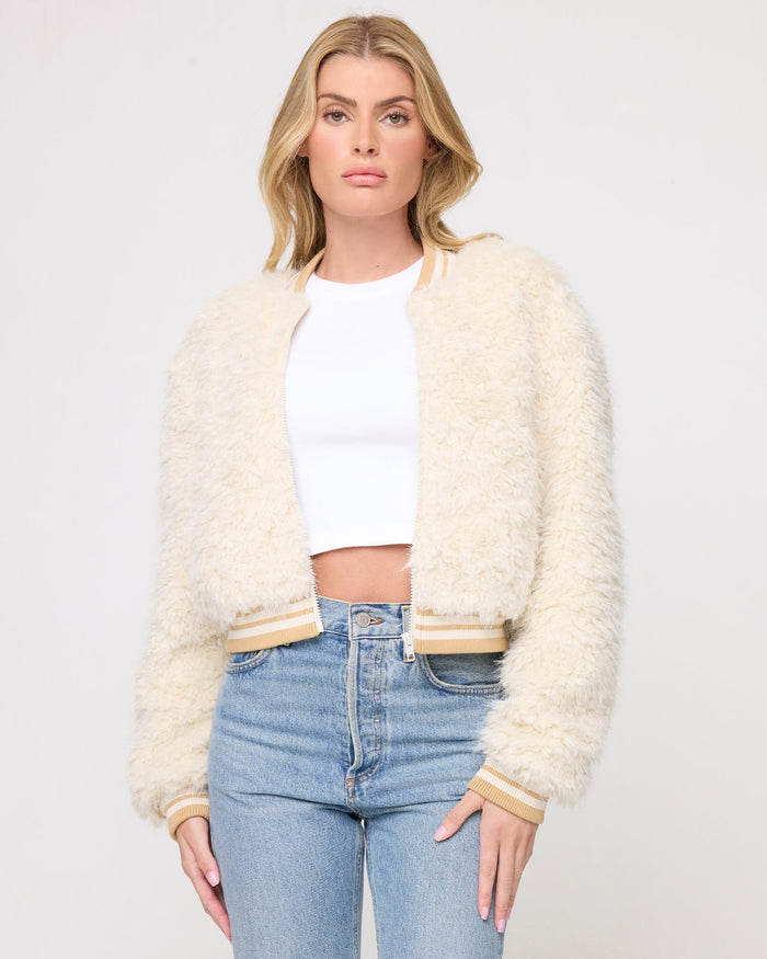 Cropped front view of a model wearing the LSPACE Valley Jacket in tan tipping, a cream shaggy teddy-bear bomber jacket with tan and cream striped ribbed collar, cuffs, and hem band, styled open over a white crop top with jeans on a gray background.