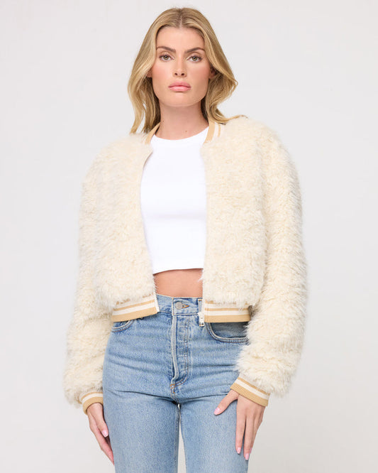Cropped front view of a model wearing the LSPACE Valley Jacket in tan tipping, a cream shaggy teddy-bear bomber jacket with tan and cream striped ribbed collar, cuffs, and hem band, styled open over a white crop top with jeans on a gray background.