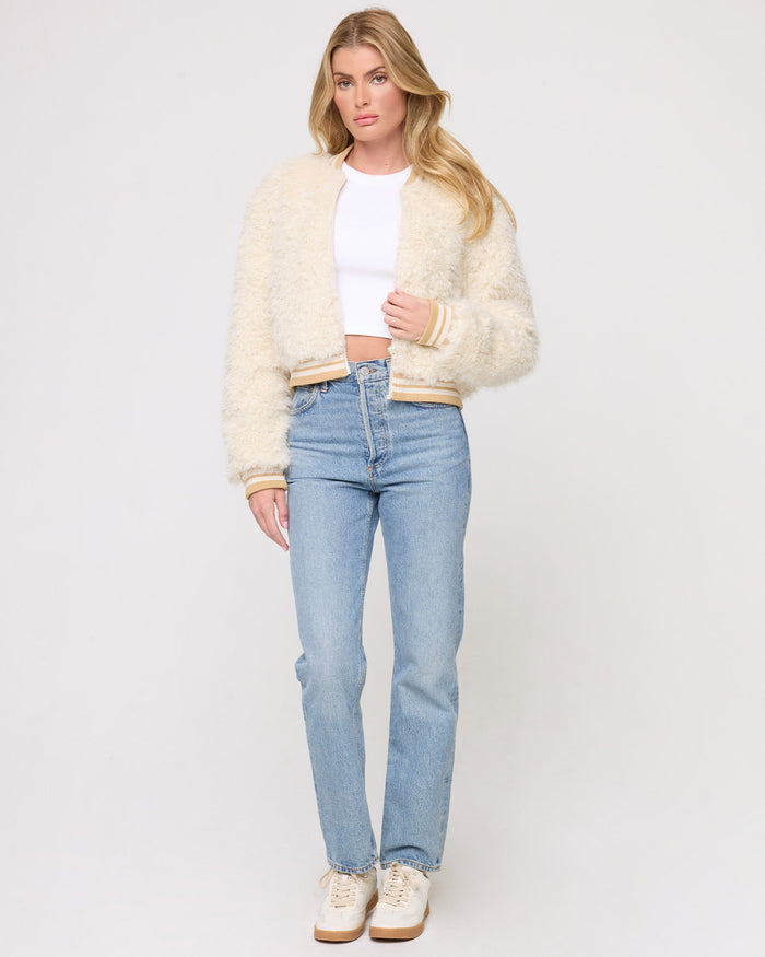 Full-length front view of a model wearing the LSPACE Valley Jacket in tan tipping, a cream shaggy bomber jacket with tan-tipped ribbed collar, cuffs, and hem, styled open over a white crop top with light blue jeans and cream sneakers on a gray background.