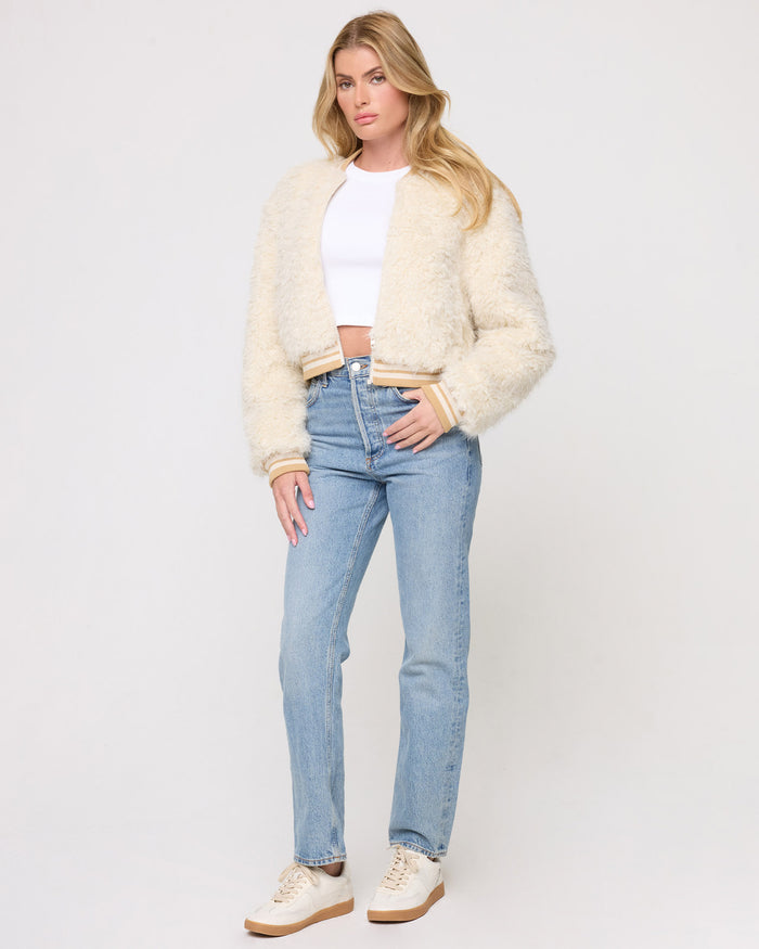 Three-quarter front view of a model wearing the LSPACE Valley Jacket in tan tipping, showing the cream shaggy texture and tan ribbed trim, styled with a white crop top, jeans, and cream sneakers on a gray background.