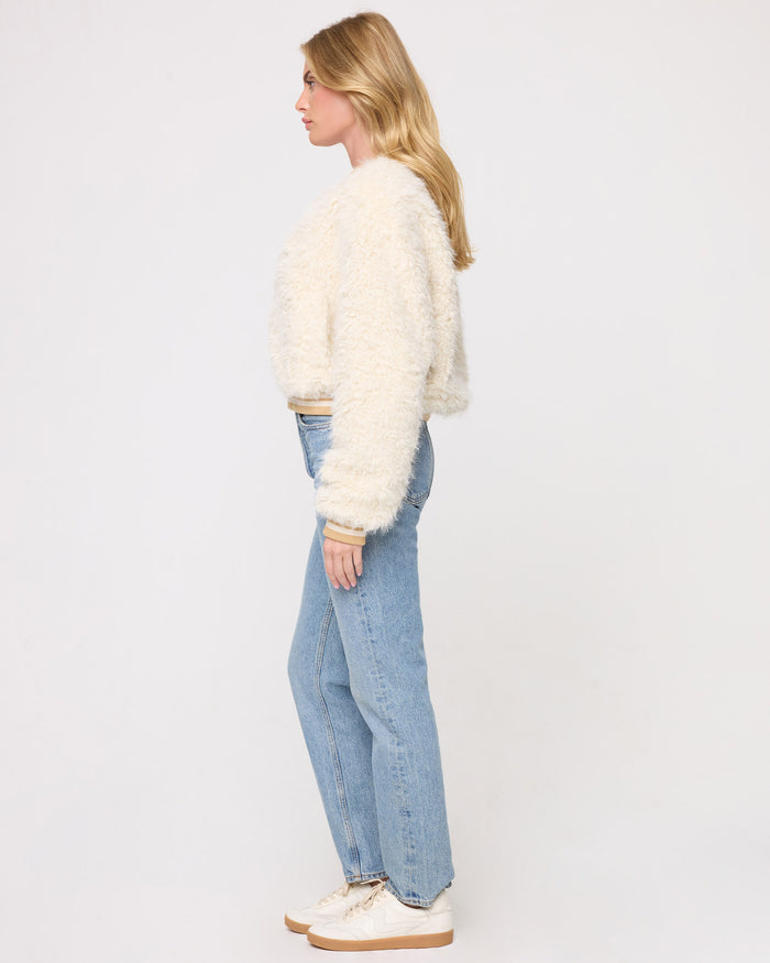 Side profile view of a model wearing the LSPACE Valley Jacket in tan tipping, showing the cropped length and voluminous shaggy cream silhouette with tan-tipped cuffs, paired with jeans and cream sneakers on a gray background.