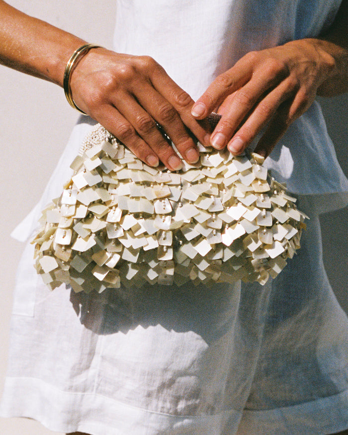 Model holding the Vienna Clutch in cream against a white linen outfit, showing the layered shell paillette texture.