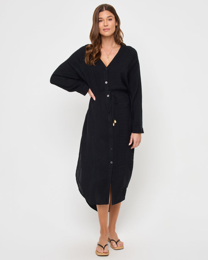 Front view of a model wearing the LSPACE Yoli Shirtdress in black, a relaxed-fit midi-length shirtdress with a V-neckline, button-front closure, long sleeves, and a drawstring waist with shell charm details.