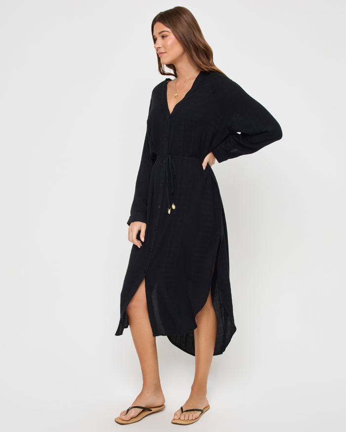 Side view of a model wearing the LSPACE Yoli Shirtdress in black, showing the side profile with the V-neckline, long dolman sleeves, drawstring waist, and high-low hem with a side slit.