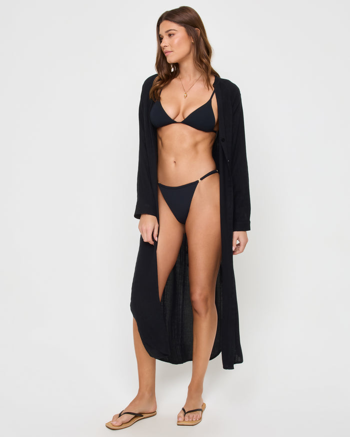 Angled front view of a model wearing the LSPACE Yoli Shirtdress in black, styled fully open as a maxi-length beach coverup over a black bikini, showing the long flowing silhouette.